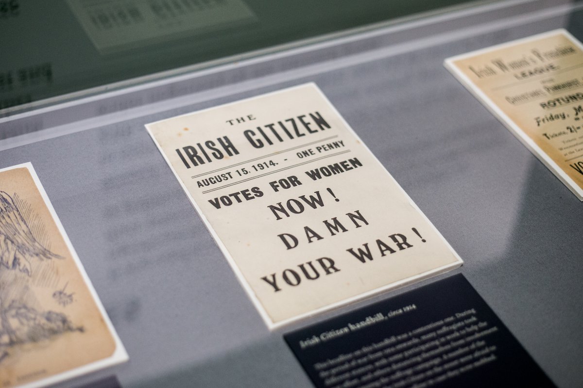 Photograph of Irish Citizen handbill, on display in an exhibition case, reading 'Votes for women now! Damn your War!'