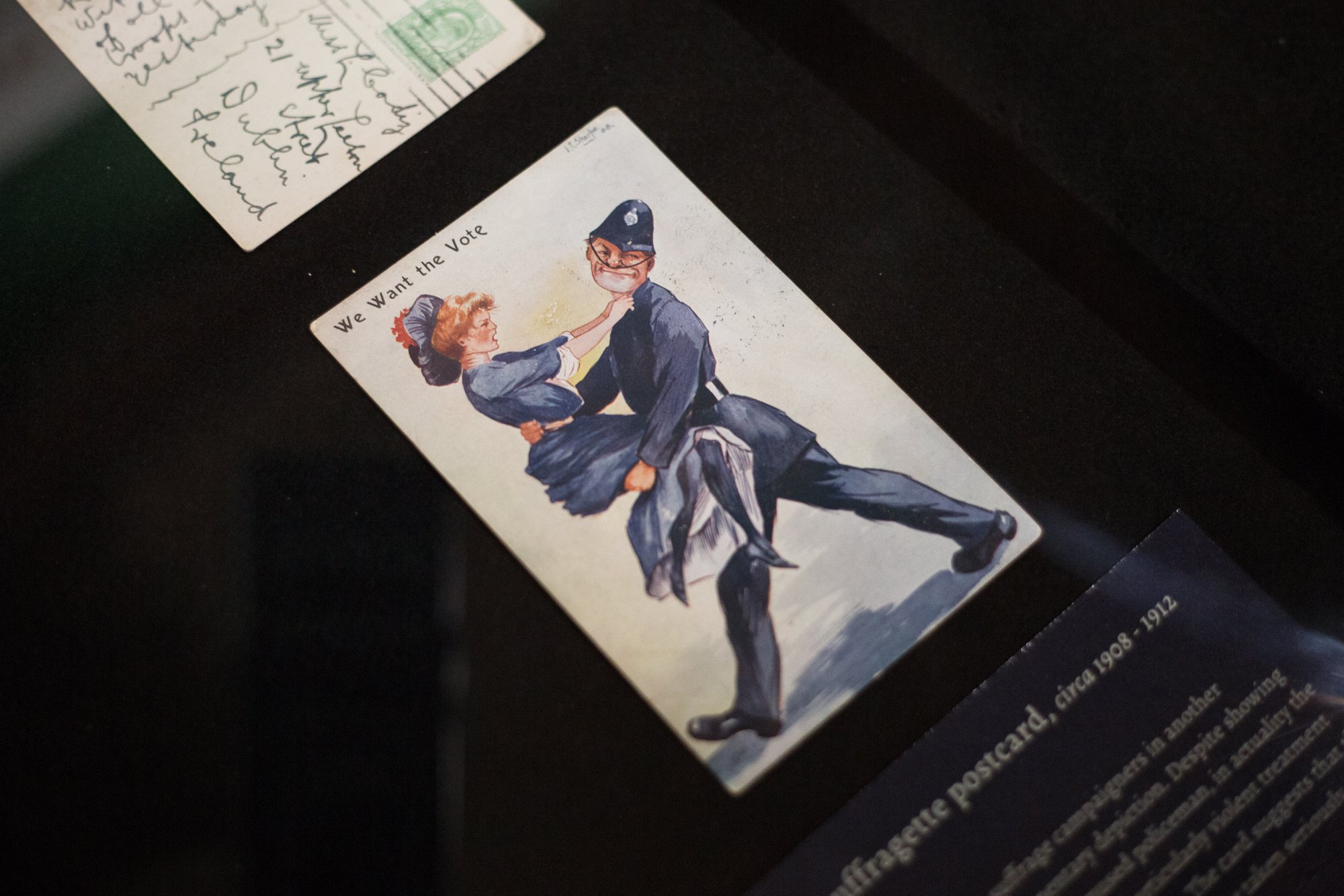 A postcard reading 'We Want the Vote' and depicting a woman being carried by a police officer, on display in an exhibition case