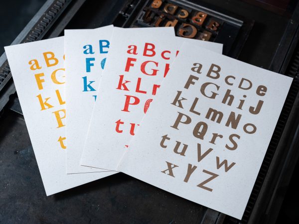 An image of four letterpress prints of the alphabet, in four different colours, resting atop the forme with which they were created