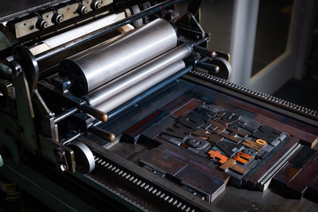 A forme containing wood type spelling out 'Now Seize that Moment' lays on the bed of a proofing press, ready to be inked up and printed.