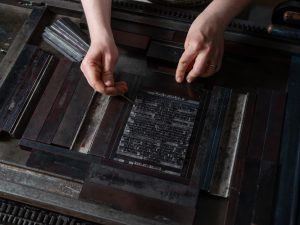 A compositor uses tools to straighten small letters of metal type in a forme.
