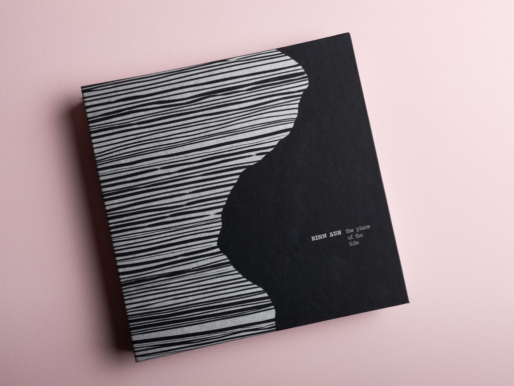 The cover view of the artist book Rinn Aun - the place of the tide. Black paper with a silver print pattern of a coast and the title text.